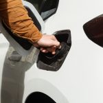A close-up of a person in a long brown denim shirt opening a white car's fuel cap on a bright, sunny day.