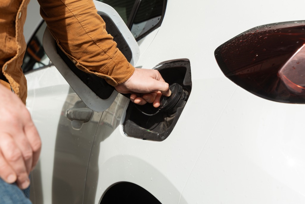 A close-up of a person in a long brown denim shirt opening a white car's fuel cap on a bright, sunny day.