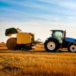 A side view of a blue tractor harvesting straw in a field with a yellow hay baler as it releases a round hay bale behind it.
