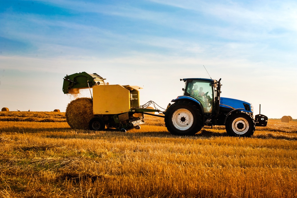 A side view of a blue tractor harvesting straw in a field with a yellow hay baler as it releases a round hay bale behind it.