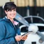 A close-up of a smiling woman holding a tablet, standing in front of a small airplane inside an aircraft hangar.