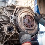 A close-up of a pair of hands with gloves grabbing the flywheel from a removed engine block of a car in a repair shop.