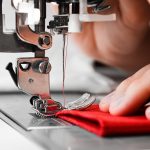 A close-up of a hand holding a folded red piece of fabric under the needle of an industrial sewing machine.