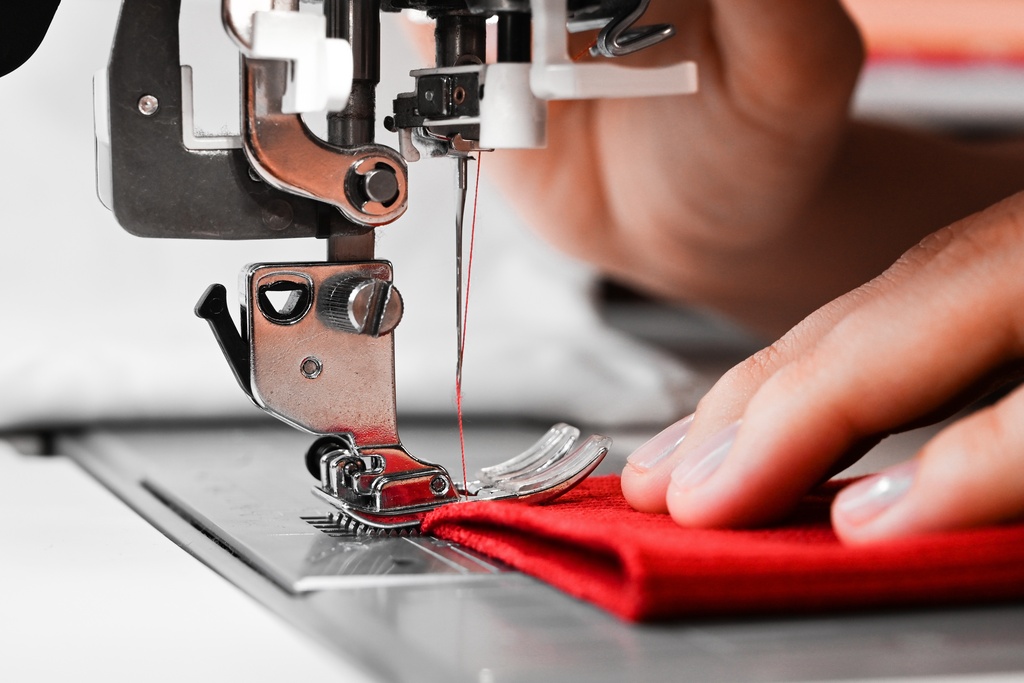 A close-up of a hand holding a folded red piece of fabric under the needle of an industrial sewing machine.