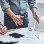 A man in a tucked in shirt is gesturing to a blueprint on a table. There is a hard hat resting on the table next to the blueprint.