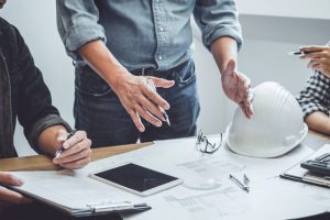 A man in a tucked in shirt is gesturing to a blueprint on a table. There is a hard hat resting on the table next to the blueprint.