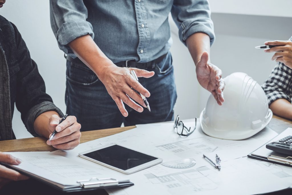 A man in a tucked in shirt is gesturing to a blueprint on a table. There is a hard hat resting on the table next to the blueprint.