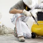 A worker in a hard hat uses a pneumatic drill attached to a yellow air compressor while breaking concrete indoors.