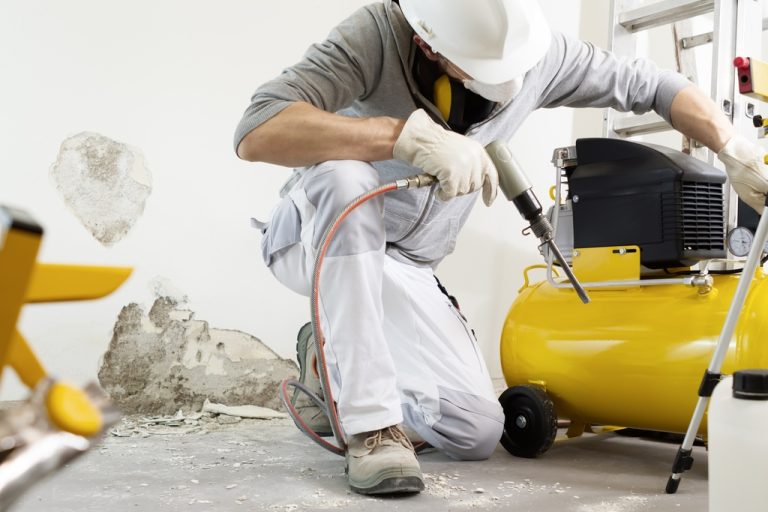 A worker in a hard hat uses a pneumatic drill attached to a yellow air compressor while breaking concrete indoors.