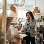 A shop owner speaking on the phone while working on a laptop behind the counter in a boutique store.
