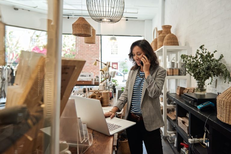 A shop owner speaking on the phone while working on a laptop behind the counter in a boutique store.