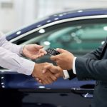 A car buyer and a suited salesperson shake hands as a key fob is passed over in front of a blue car at a dealership.