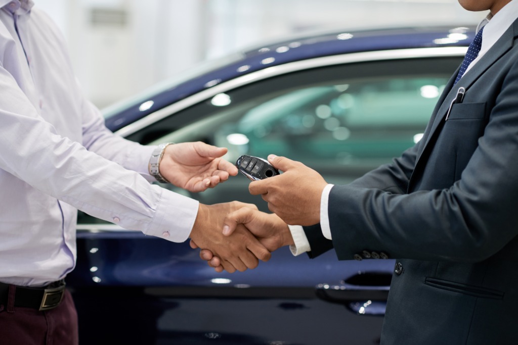 A car buyer and a suited salesperson shake hands as a key fob is passed over in front of a blue car at a dealership.