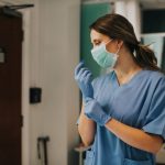 A woman in blue medical scrubs is pulling a blue glove over her hand. She is also wearing a face mask.