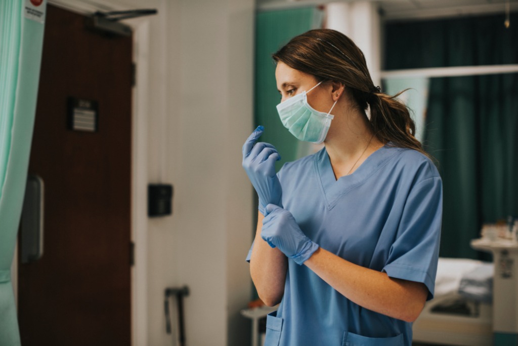 A woman in blue medical scrubs is pulling a blue glove over her hand. She is also wearing a face mask.