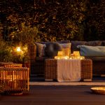 A backyard patio lounge with a wicker sofa, glowing lanterns and candles at dusk, surrounded by lush plants and warm lighting.