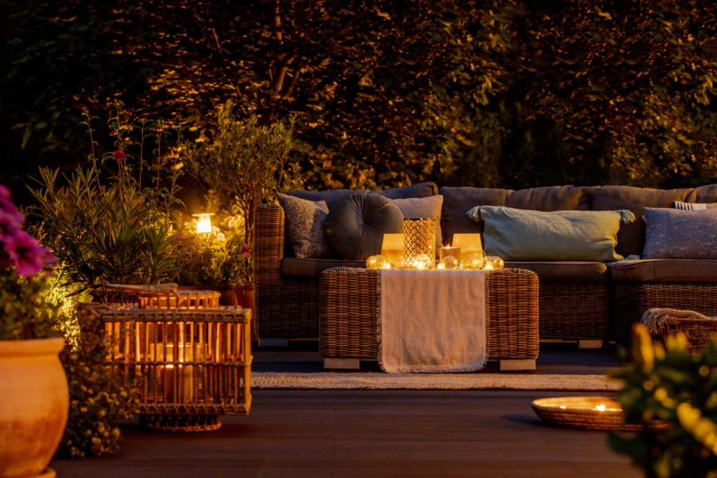 A backyard patio lounge with a wicker sofa, glowing lanterns and candles at dusk, surrounded by lush plants and warm lighting.
