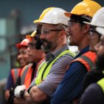 A group of employees stand together in a line with hard hats and high-vis safety vests on. Some have safety glasses on.