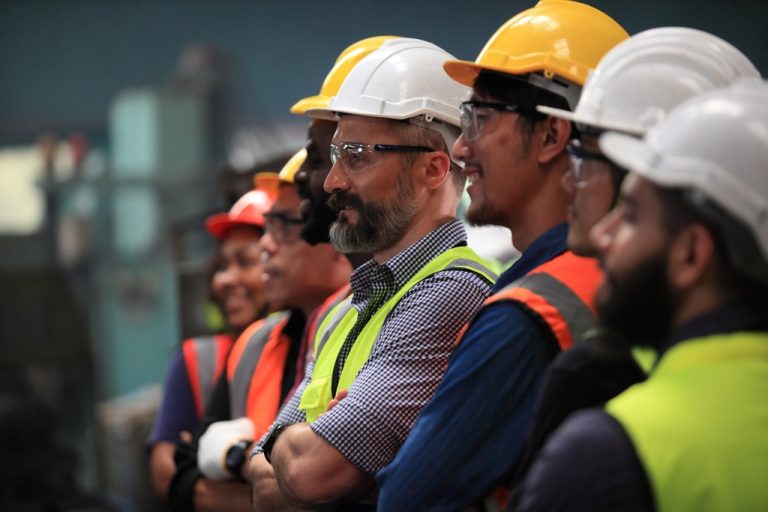 A group of employees stand together in a line with hard hats and high-vis safety vests on. Some have safety glasses on.