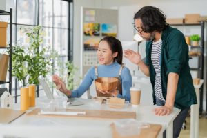 Two people gather around a table discussing something. Various types of packaging surround the table in front of them.