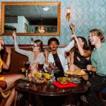 A group of friends seated at a restaurant table all holding their adult beverages in the air for a "cheers!"