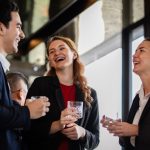 A group of coworkers standing and talking together at a social gathering with drinks and lights in the background.