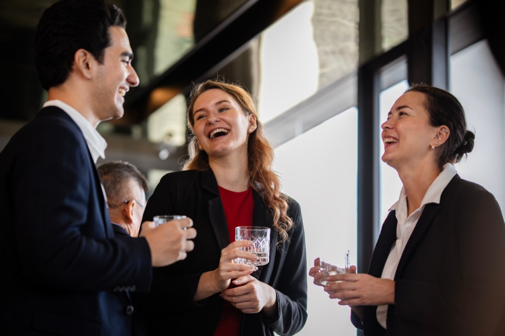 A group of coworkers standing and talking together at a social gathering with drinks and lights in the background.