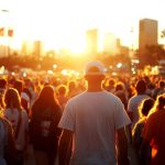 An outdoor event features hundreds of people walking down a street. In the background, the sun sets behind a city skyline.