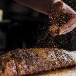A hand sprinkles coarse seasoning over a thick raw steak on a cutting board, spices scattering in the air above the steak.