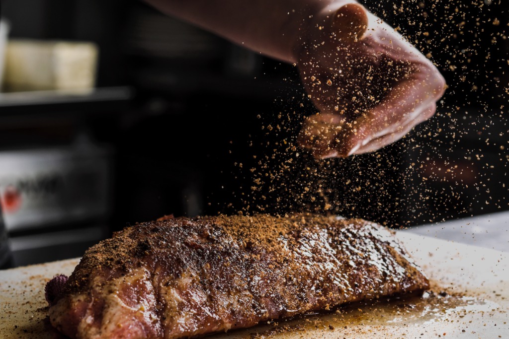 A hand sprinkles coarse seasoning over a thick raw steak on a cutting board, spices scattering in the air above the steak.