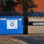 Two large commercial waste hoppers sit side-by-side in the business parking lot. One is for recycle and one is for trash.