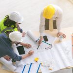 Three construction workers wearing hard hats and leaning over a table. Several blueprints are on the table.