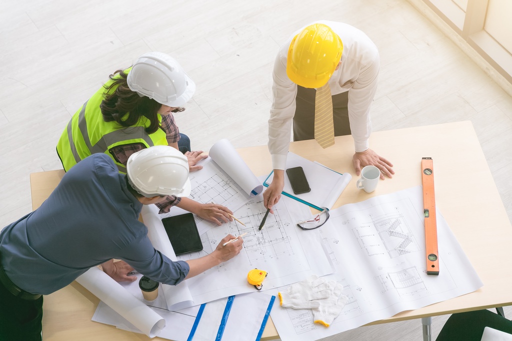 Three construction workers wearing hard hats and leaning over a table. Several blueprints are on the table.
