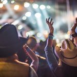 The crowd at a live concert wearing cowboy hats with their hands raised toward the bright stage lights.