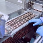 Gloved hands guiding rows of capsules on a pharmaceutical production line, with machinery filling and organizing pills.