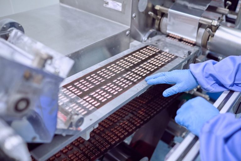 Gloved hands guiding rows of capsules on a pharmaceutical production line, with machinery filling and organizing pills.