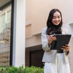 A smiling woman wearing a tan blazer and white pants is holding a tablet with a black case, standing outside a building.