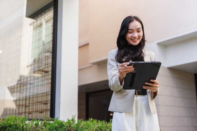 A smiling woman wearing a tan blazer and white pants is holding a tablet with a black case, standing outside a building.