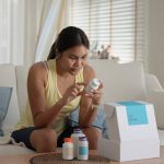 A young woman sits on a cream-colored couch taking bottled products out of white cardboard box.