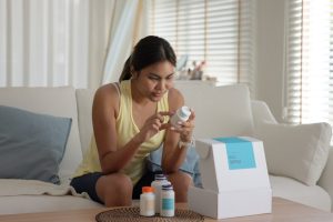 A young woman sits on a cream-colored couch taking bottled products out of white cardboard box.