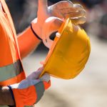 A close-up of a person wearing an orange safety vest and orange and white work gloves while holding a yellow hard hat.