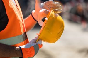 A close-up of a person wearing an orange safety vest and orange and white work gloves while holding a yellow hard hat.