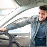 A man leans into a car with the side passenger door open. His hand rests on the dash and he smiles.