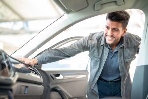A man leans into a car with the side passenger door open. His hand rests on the dash and he smiles.
