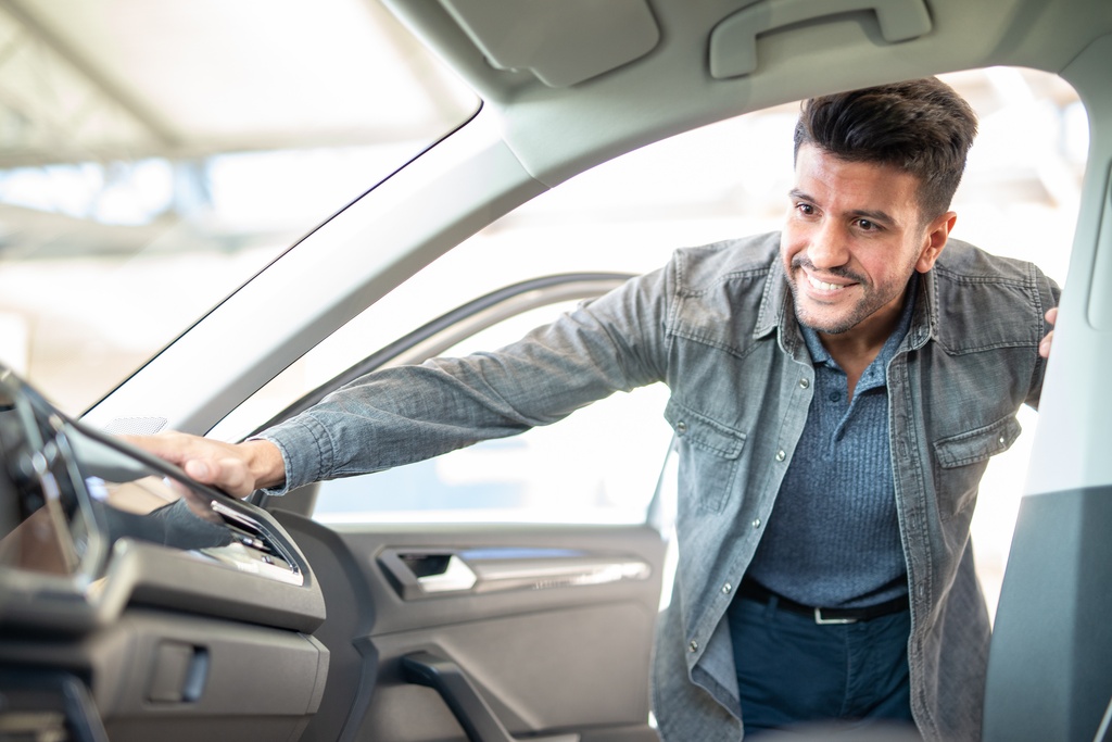 A man leans into a car with the side passenger door open. His hand rests on the dash and he smiles.
