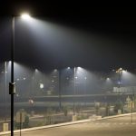 An empty parking lot at night with bright light poles, hazy air, small trees, road markings, and a speed limit sign.