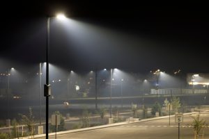 An empty parking lot at night with bright light poles, hazy air, small trees, road markings, and a speed limit sign.