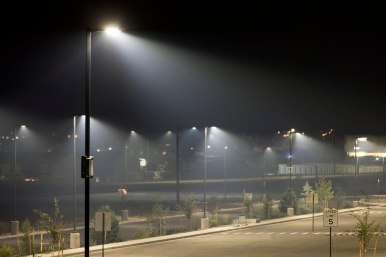 An empty parking lot at night with bright light poles, hazy air, small trees, road markings, and a speed limit sign.