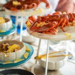 A buffet table at an event set up for guests to graze. There's a large lobster sitting on a rack and small oyster dishes.