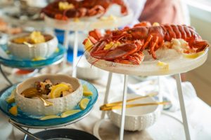 A buffet table at an event set up for guests to graze. There's a large lobster sitting on a rack and small oyster dishes.